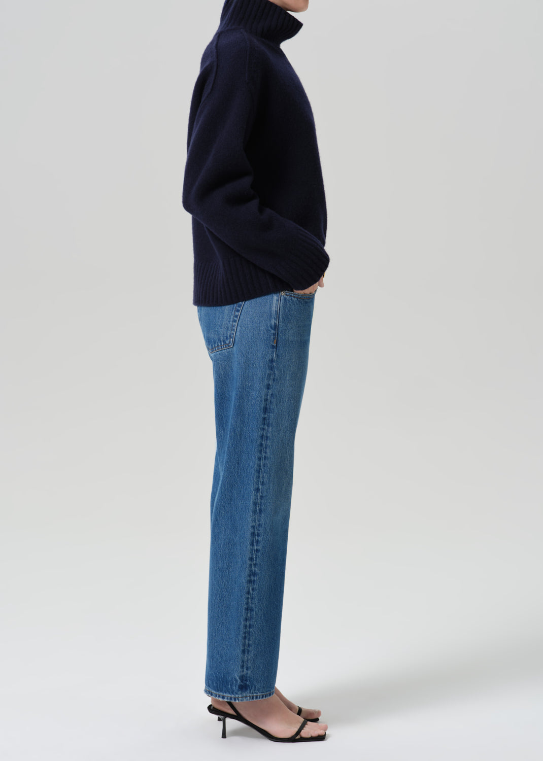A side-profile studio shot of a model wearing a dark navy blue turtleneck sweater, blue denim jeans, and black strappy high-heeled sandals against a grey background.
