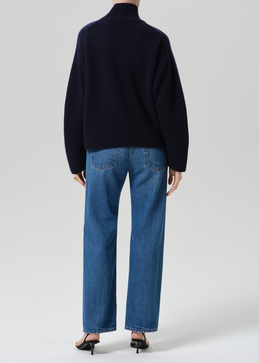 A model shown from the back wearing a dark navy blue turtleneck sweater and blue wide-leg jeans, paired with black heels, against a grey background.