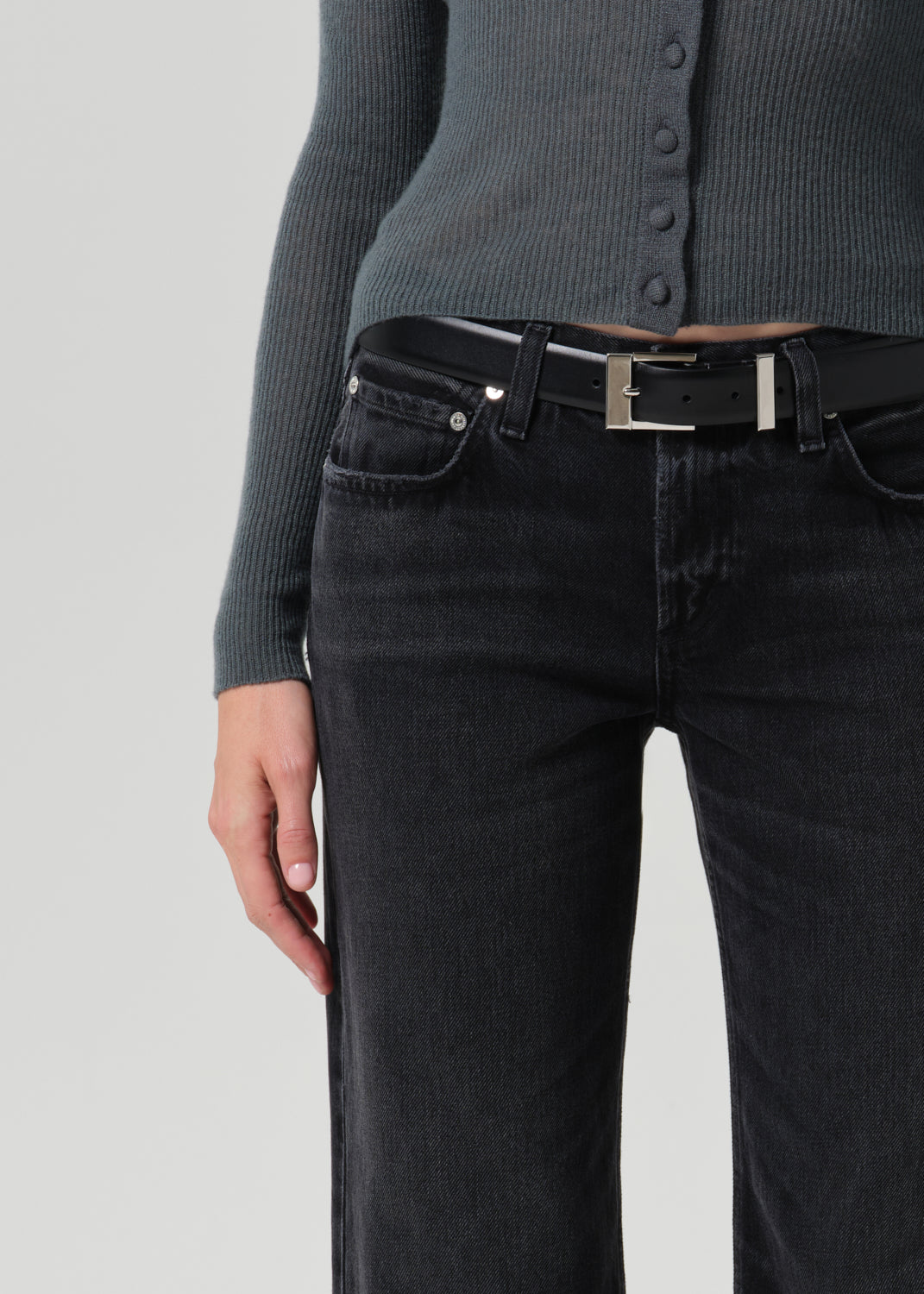 Close-up of a person wearing dark charcoal grey jeans with a black belt featuring a silver buckle, paired with a ribbed grey cardigan.