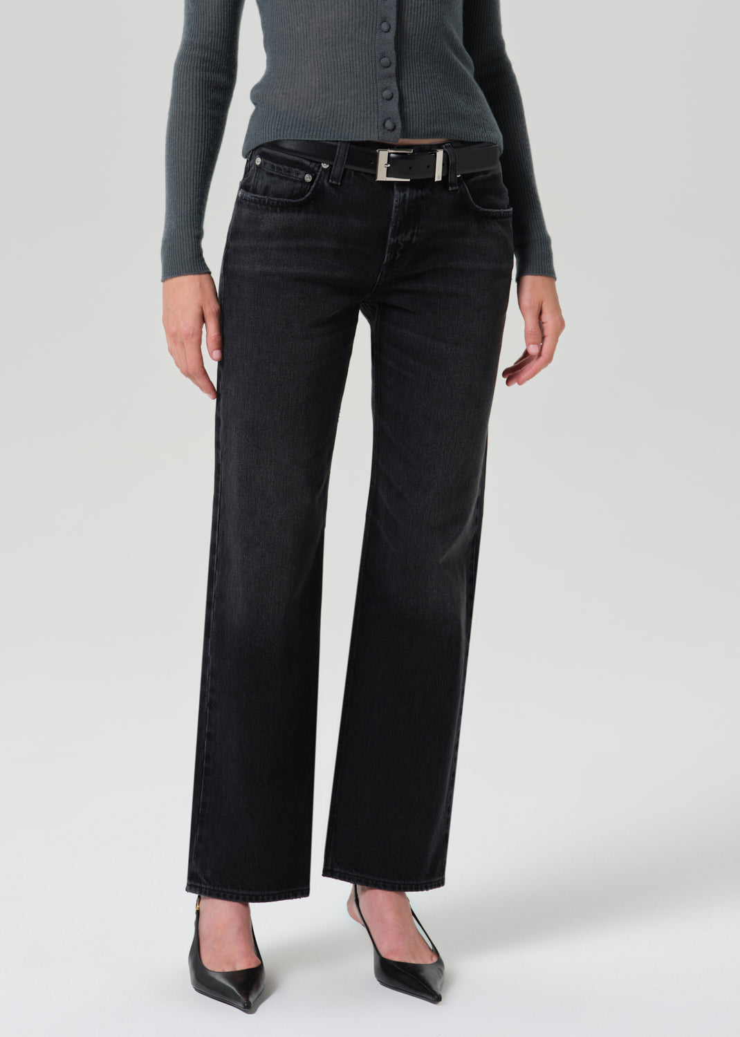 A model wearing a pair of black wide-leg jeans with a silver buckle belt, styled with a dark grey button-up cardigan and black pointed-toe heels against a grey background.