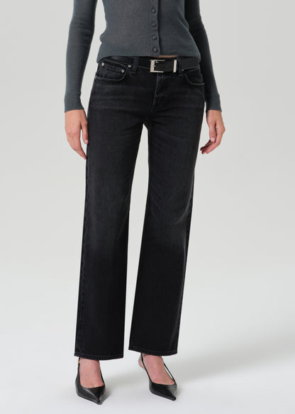 A model wearing a pair of black wide-leg jeans with a silver buckle belt, styled with a dark grey button-up cardigan and black pointed-toe heels against a grey background.