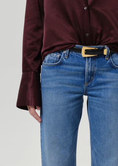 Close-up of a person wearing blue jeans with a black belt featuring a gold rectangular buckle, paired with a dark burgundy satin blouse.