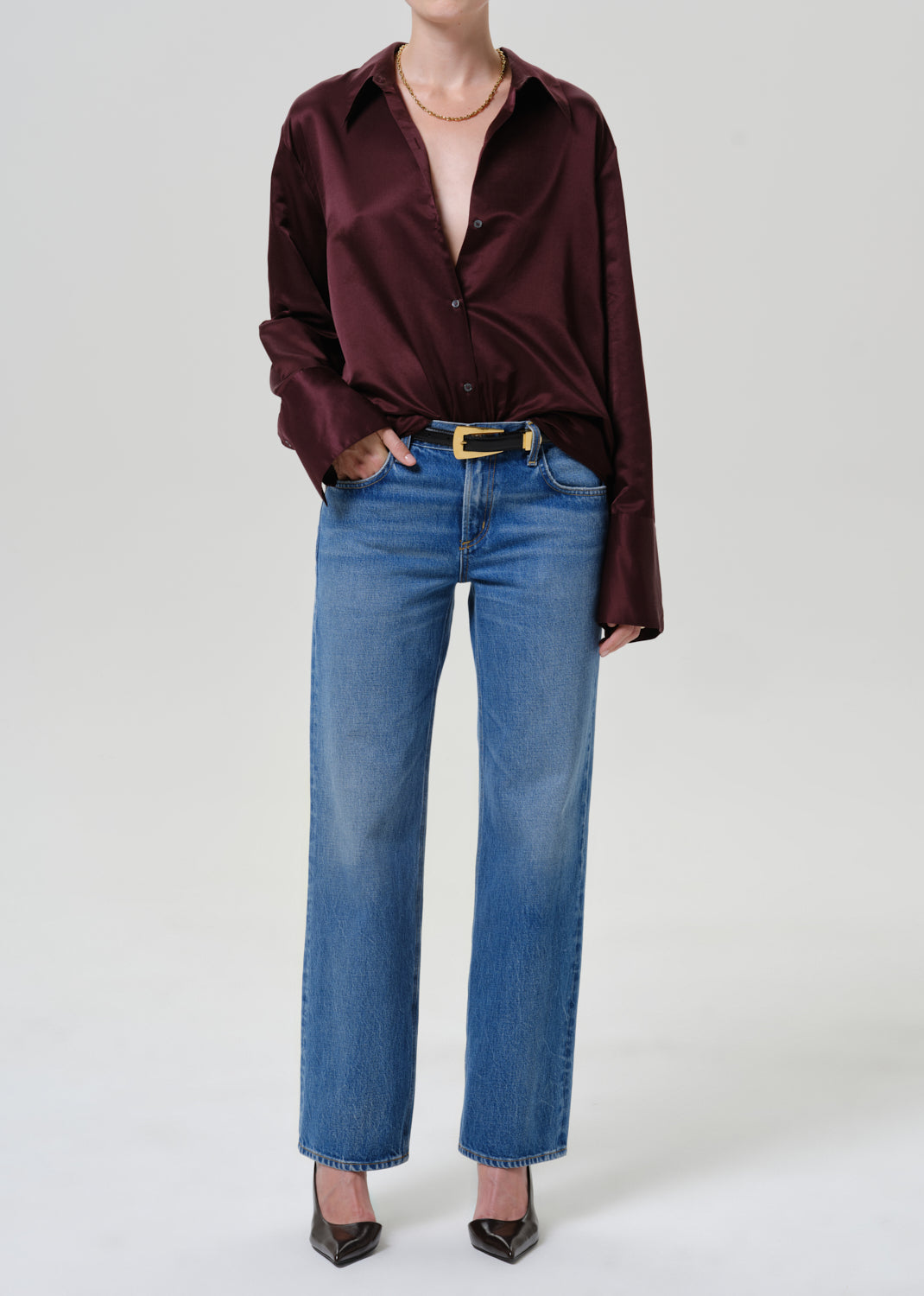 A model wearing a dark burgundy satin button-down shirt, blue straight-leg jeans, a gold belt, and black pointed-toe heels, photographed against a neutral light gray studio background.