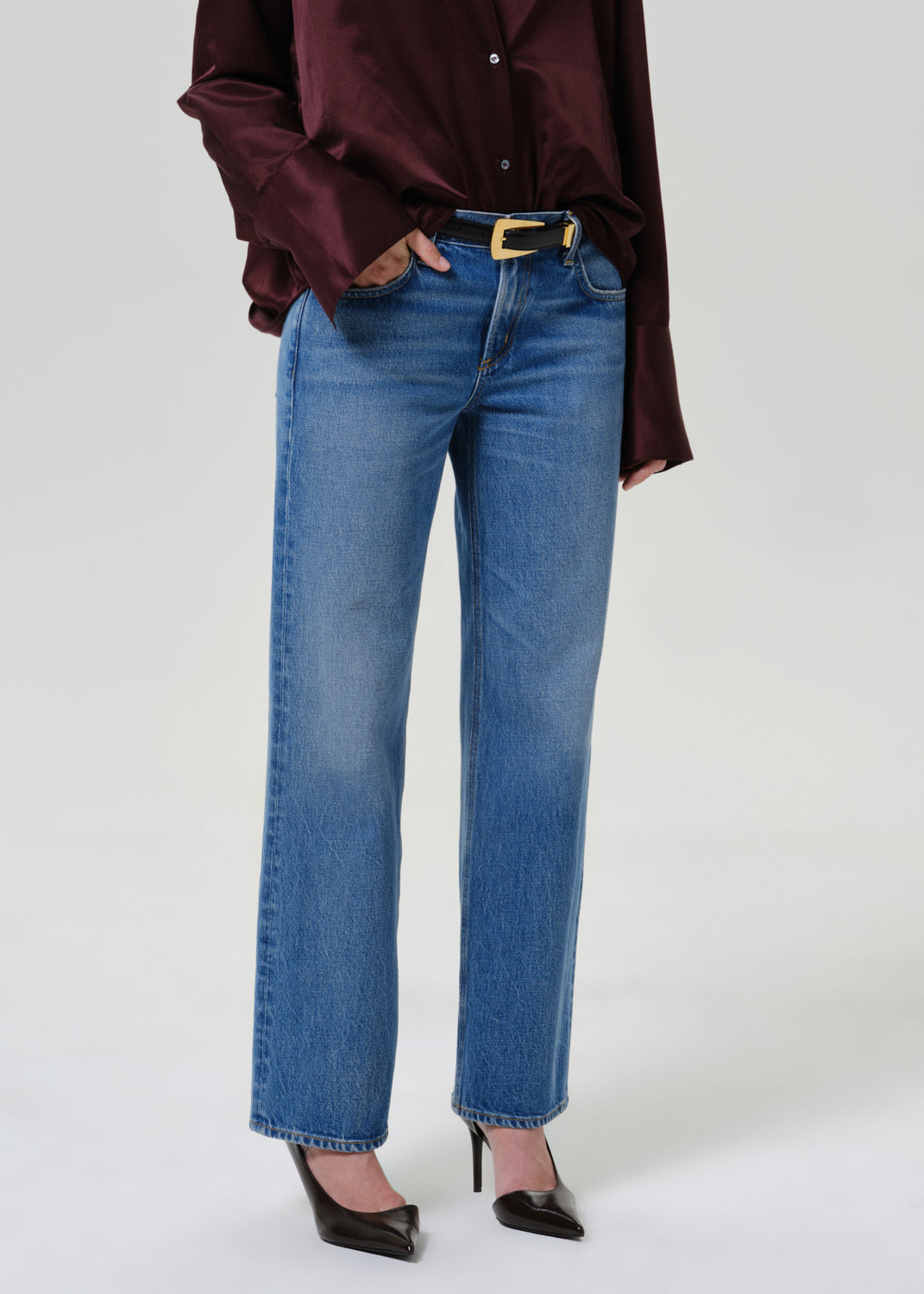 A model wearing a pair of medium-wash blue straight-leg jeans with a gold-tone belt buckle, styled with a dark burgundy satin shirt and black pointed-toe heels against a light gray studio background.
