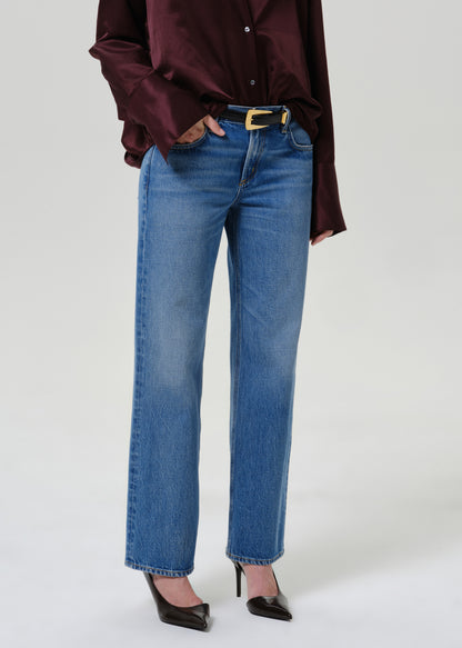 A model wearing a pair of medium-wash blue straight-leg jeans with a gold-tone belt buckle, styled with a dark burgundy satin shirt and black pointed-toe heels against a light gray studio background.