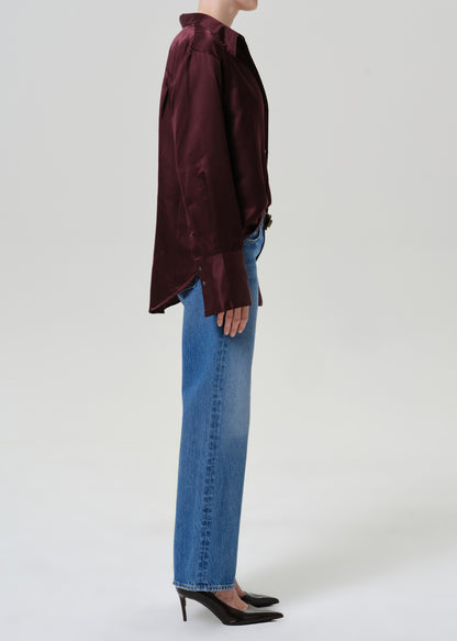 A side-profile studio shot of a model wearing a dark burgundy satin long-sleeve blouse, blue jeans, and black pointed-toe heels against a light gray background.