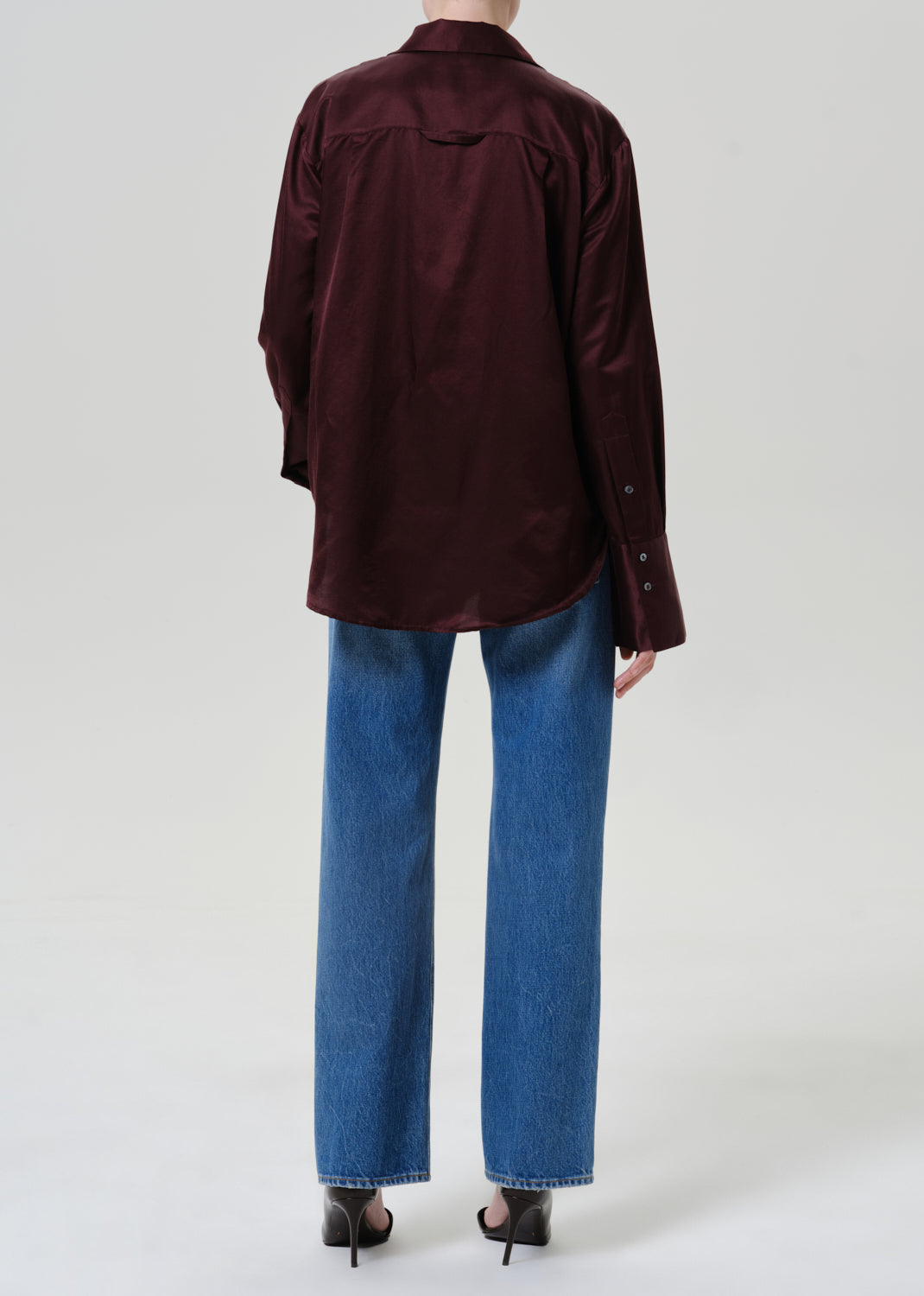 A model shown from the back wearing a dark burgundy satin button-up shirt, blue wide-leg jeans, and black heels against a light gray studio background.