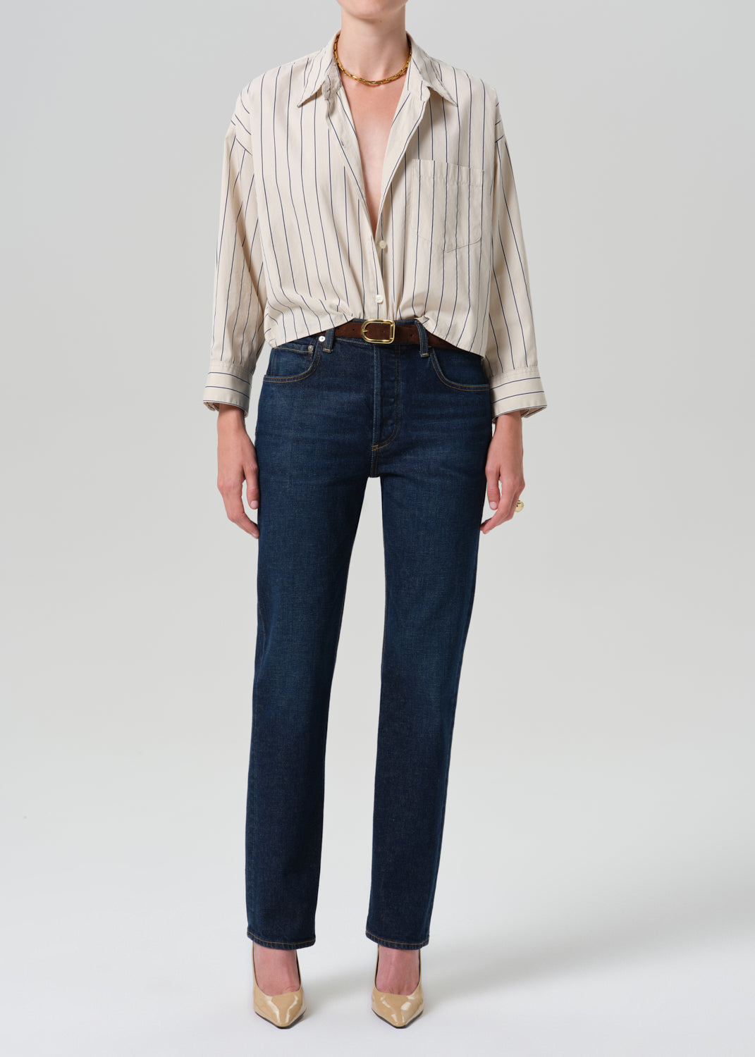 A model wearing dark blue straight-leg jeans, a cream and beige vertically striped button-down shirt, a brown leather belt, and gold pointed-toe heels, photographed against a grey background.