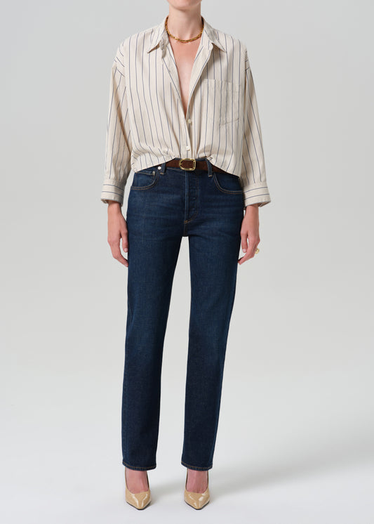 A model wearing dark blue straight-leg jeans, a cream and beige vertically striped button-down shirt, a brown leather belt, and gold pointed-toe heels, photographed against a grey background.