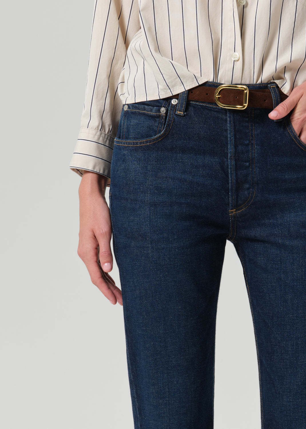 Close-up of a person wearing dark blue denim jeans with a brown leather belt featuring a gold buckle, paired with a cream pinstriped shirt.