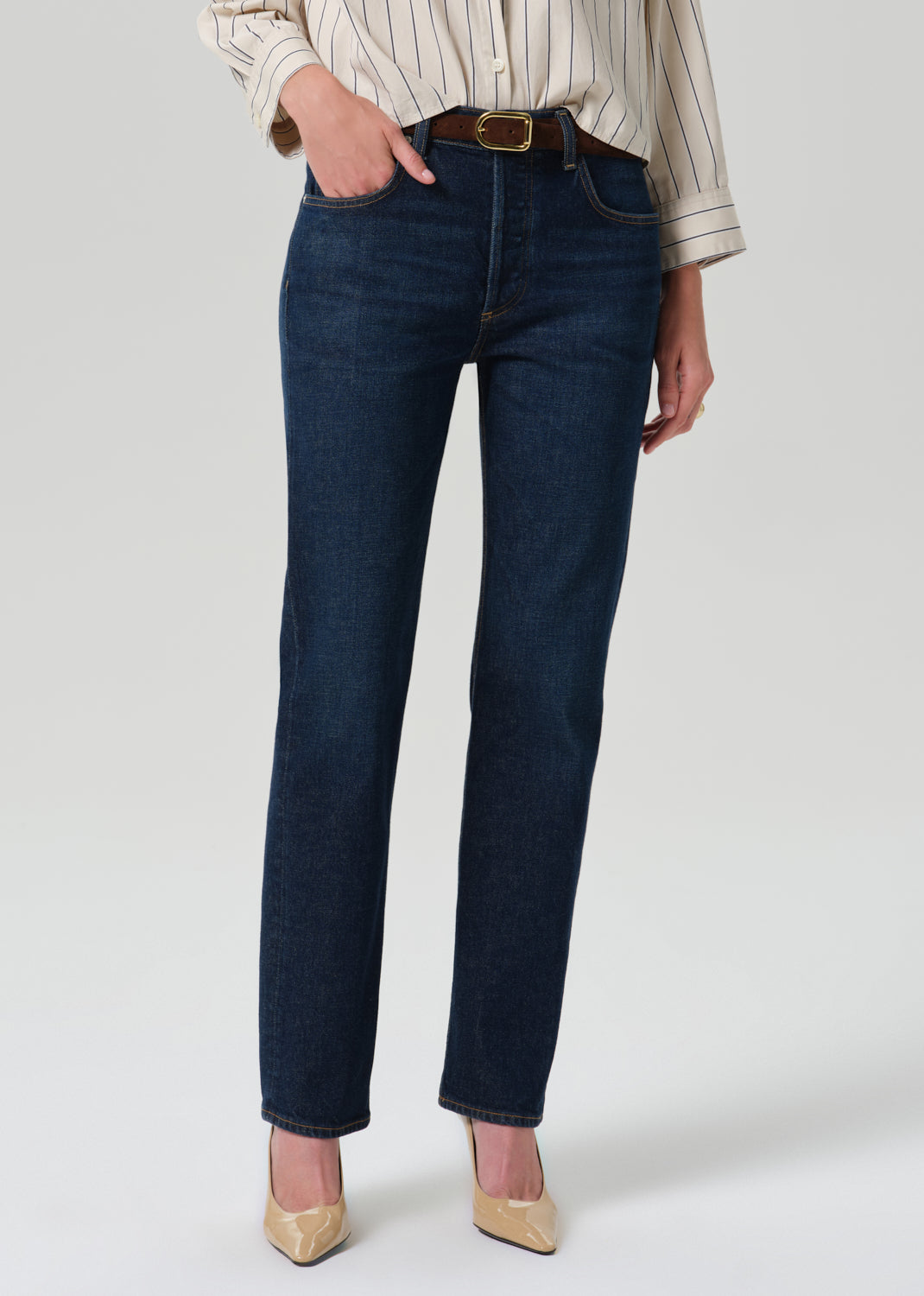 A person wearing dark blue straight-leg jeans with a brown belt, paired with a cream and brown striped shirt and beige pointed-toe heels, shown from the waist down on a grey  background.