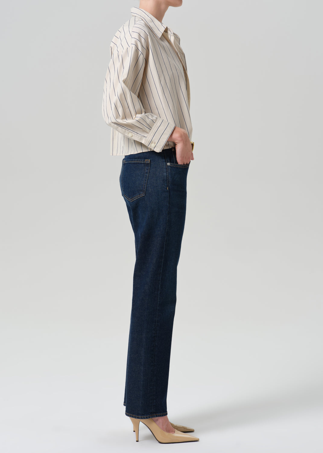 A model shown from the side wearing dark blue straight-leg jeans, a cream and brown vertically striped button-down shirt, and beige high-heeled pumps against a grey background.