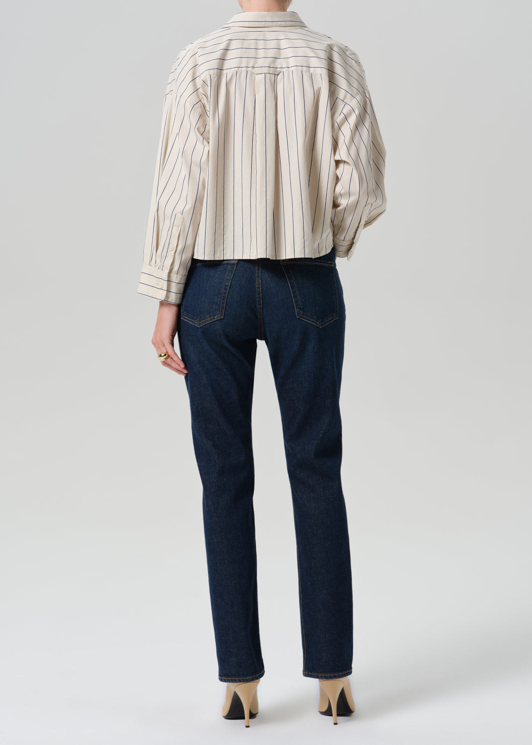 Rear view of a model wearing a cream and beige vertically striped button-up shirt and dark blue straight-leg jeans, paired with beige high heels, against a grey background.