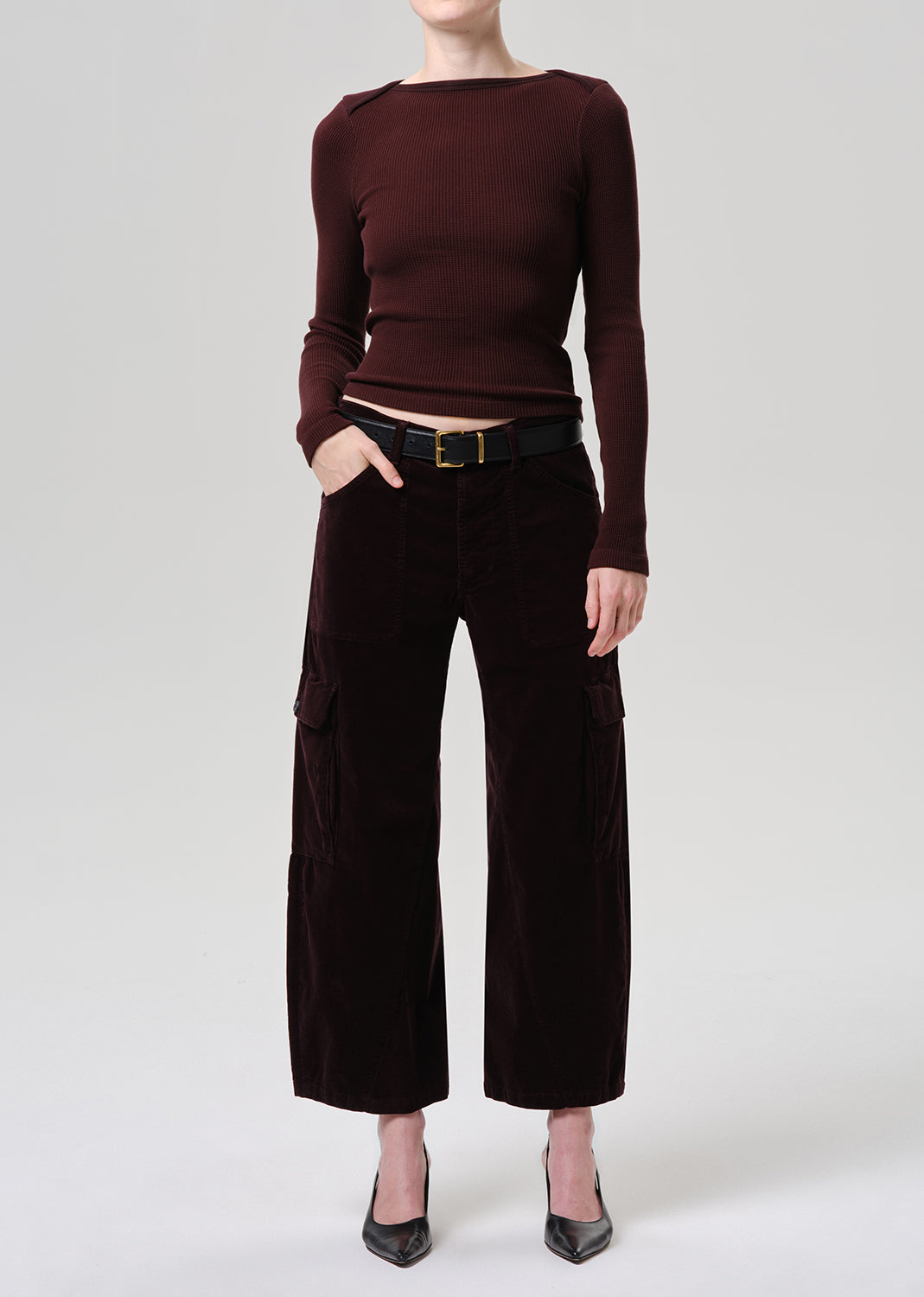 A model wearing a dark burgundy long-sleeve knit top and dark brown wide-leg velvet pants with a black belt, standing against a grey background.