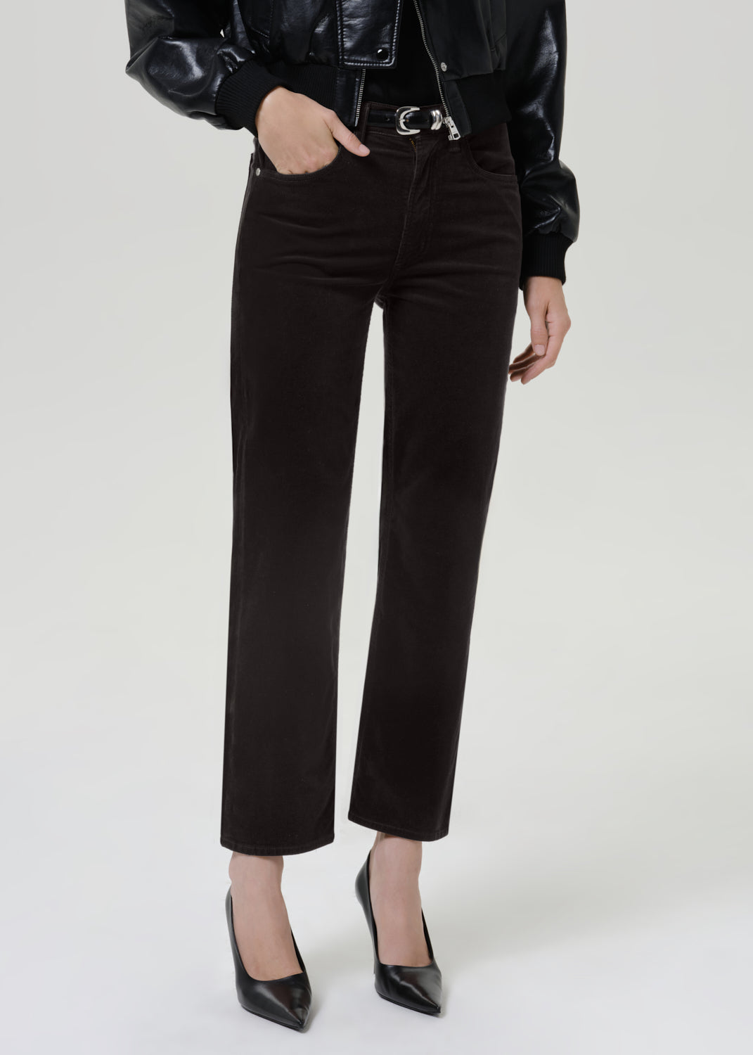 A model wearing high-waisted, straight-leg gray brown corduroy pants with a silver-tone belt, paired with a black leather jacket and black pointed-toe heels, against a light gray studio background.