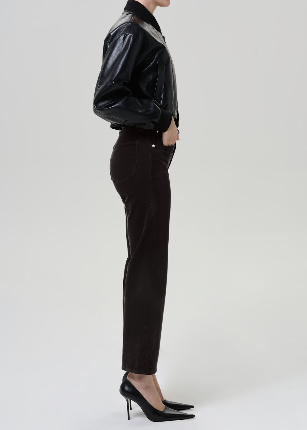 A side-profile studio shot of a model wearing a black glossy leather jacket, gray brown corduroy trousers, and black pointed-toe stiletto heels against a light gray background.