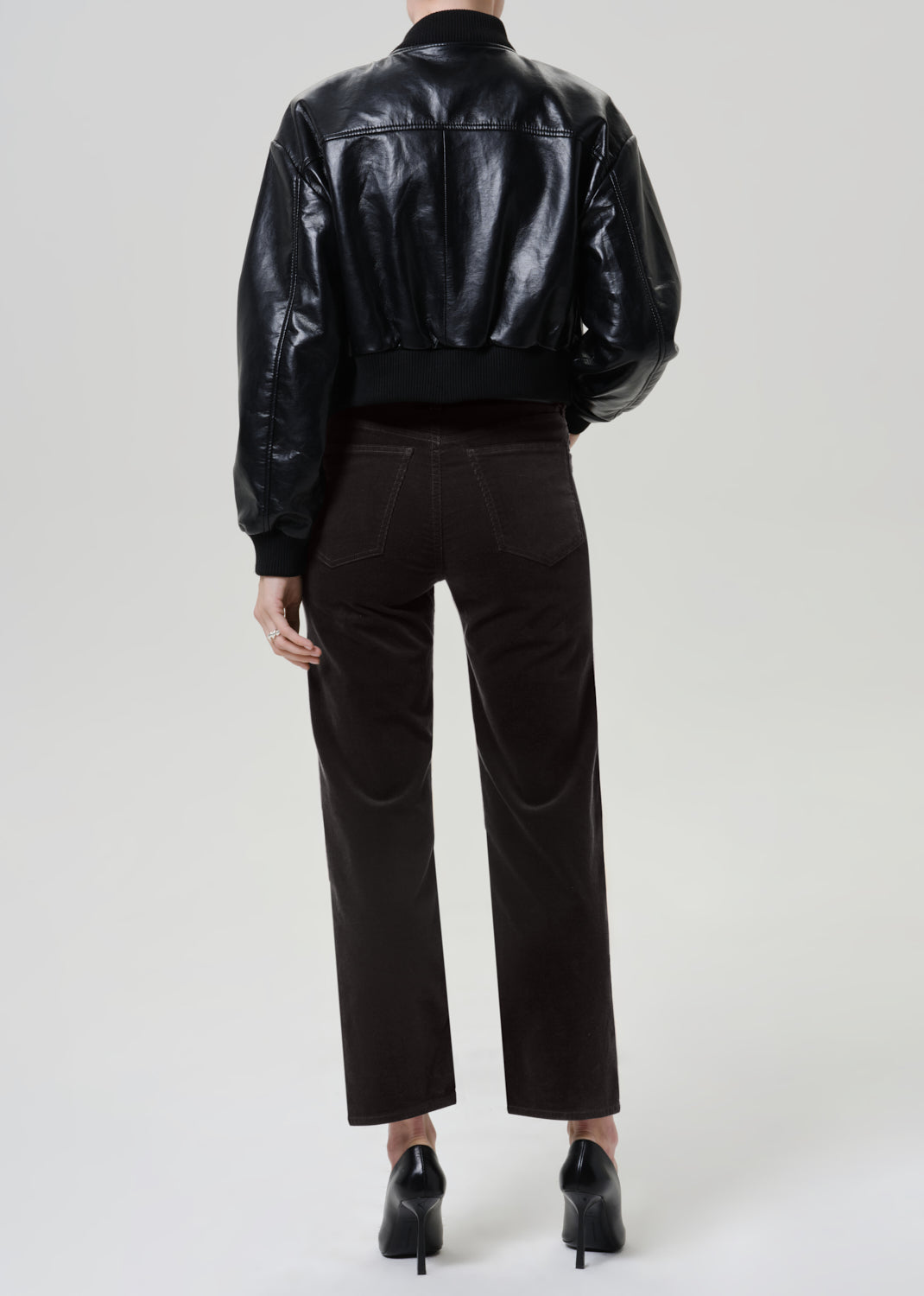 A model shown from the back wearing a shiny black leather cropped jacket, dark gray brown trousers, and black pointed-toe heels against a light gray studio background.