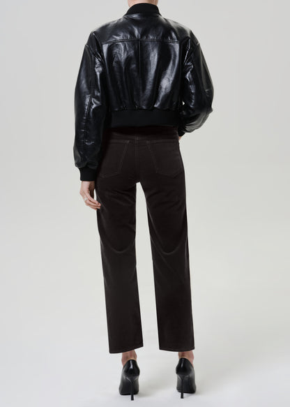 A model shown from the back wearing a shiny black leather cropped jacket, dark gray brown trousers, and black pointed-toe heels against a light gray studio background.