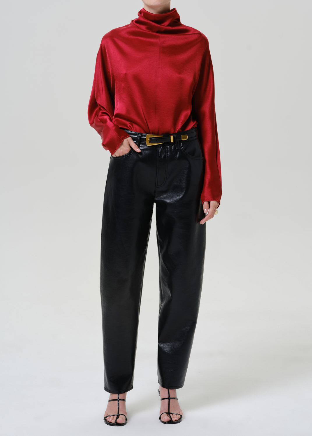 Woman wearing black leather pants with a red satin draped neck top on a grey background.