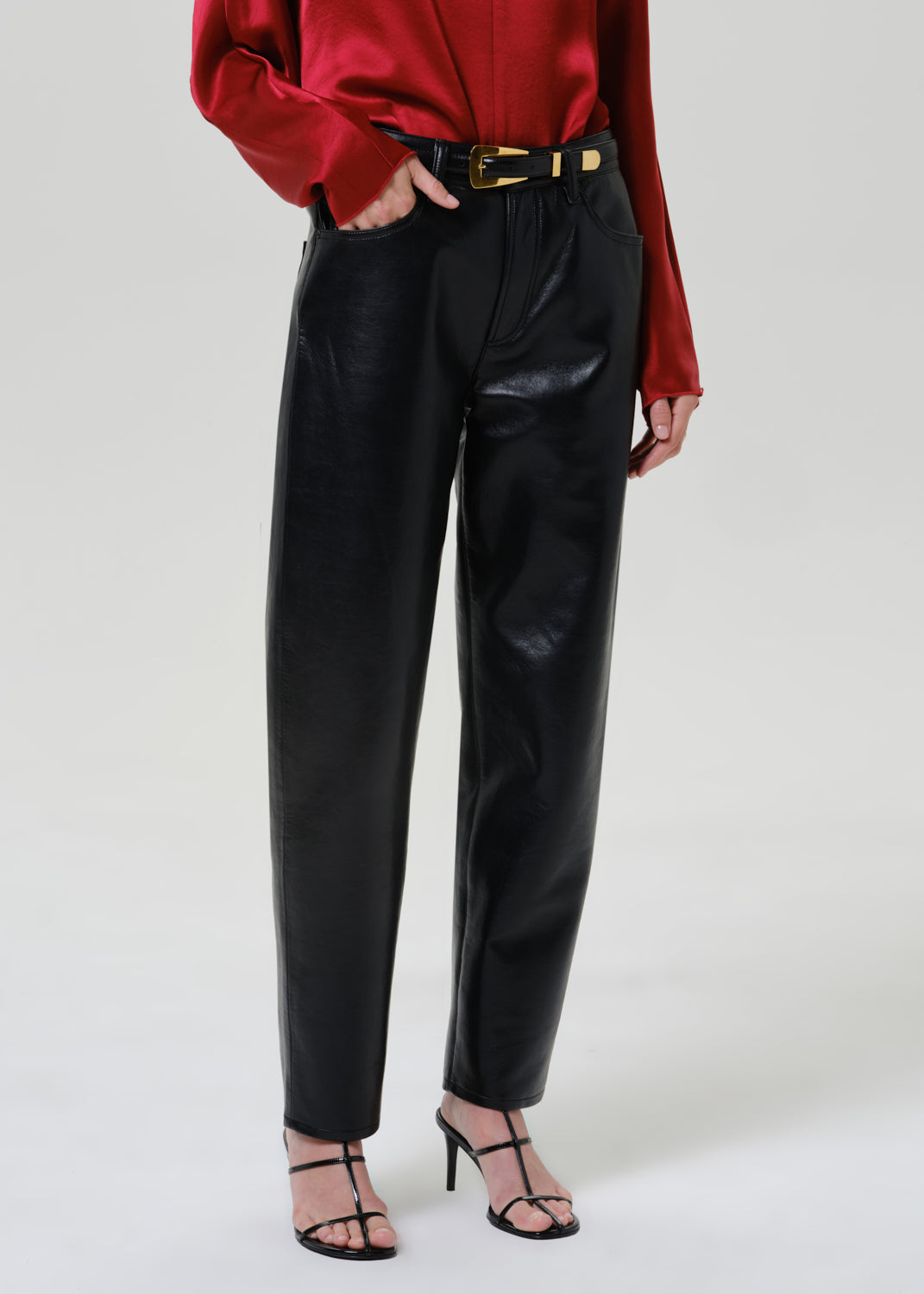 Woman wearing black leather pants with a red satin draped neck top on a grey background.