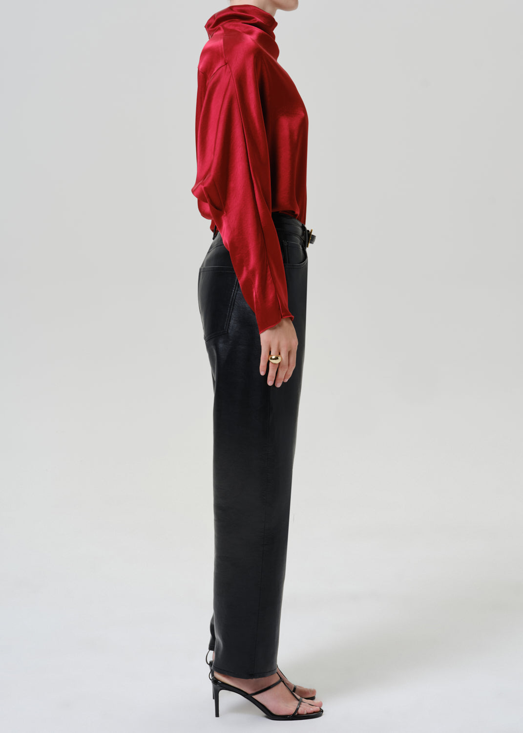 Woman wearing black leather pants with a red satin draped neck top viewed from the side on a grey background.