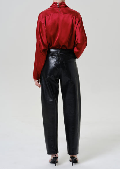 Woman wearing black leather pants with a red satin draped neck top viewed from the back on a grey background.