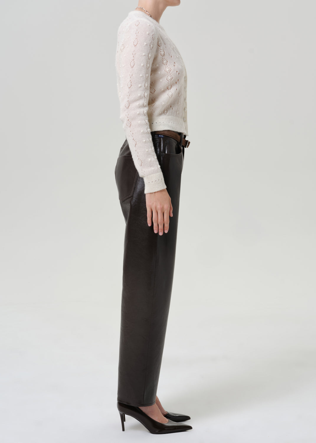Woman wearing black leather pants with an ivory button-front cardigan viewed from the side on a grey background.