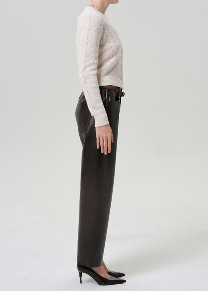 Woman wearing black leather pants with an ivory button-front cardigan viewed from the side on a grey background.