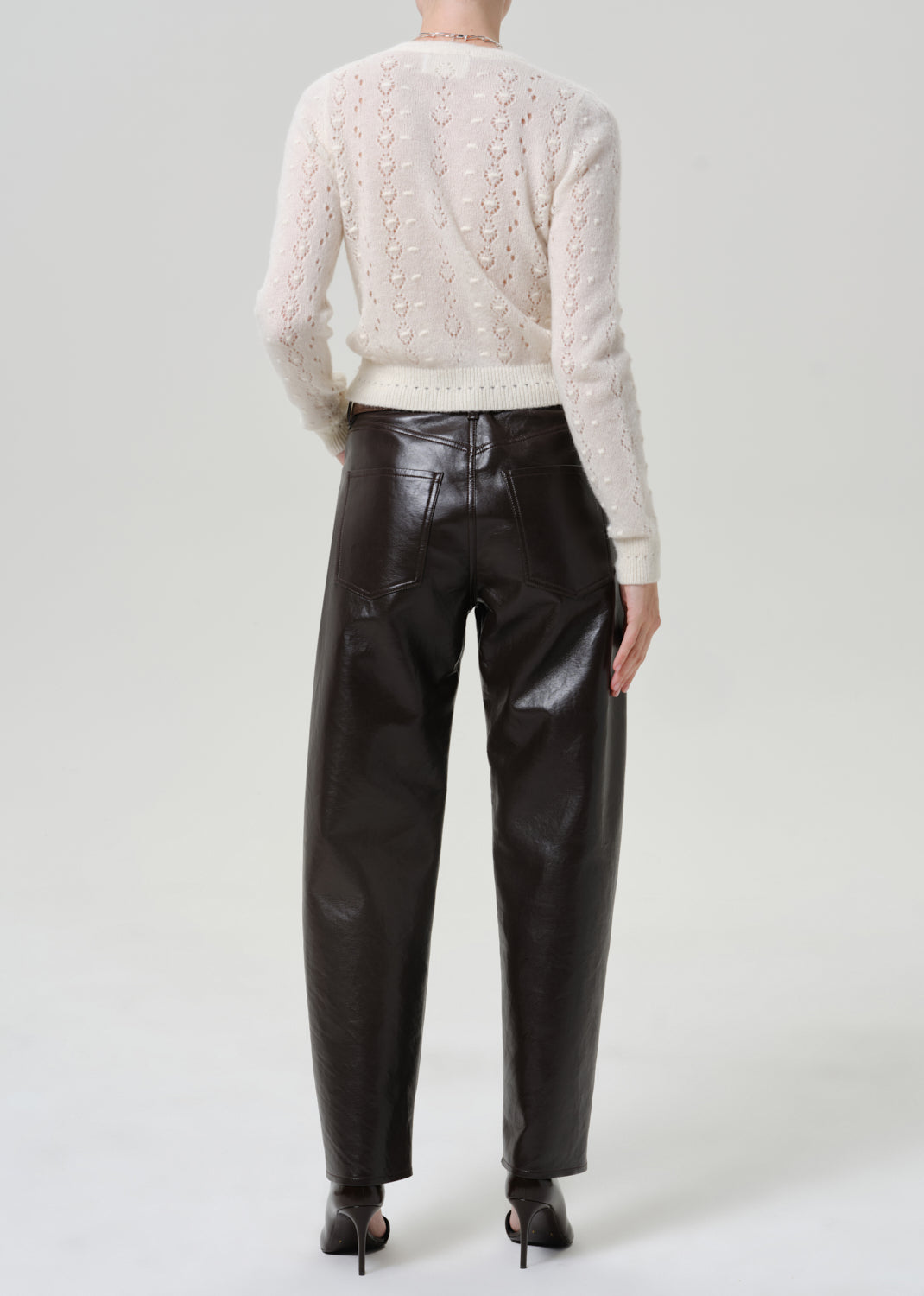 Woman wearing black leather pants with an ivory button-front cardigan viewed from the back on a grey background.