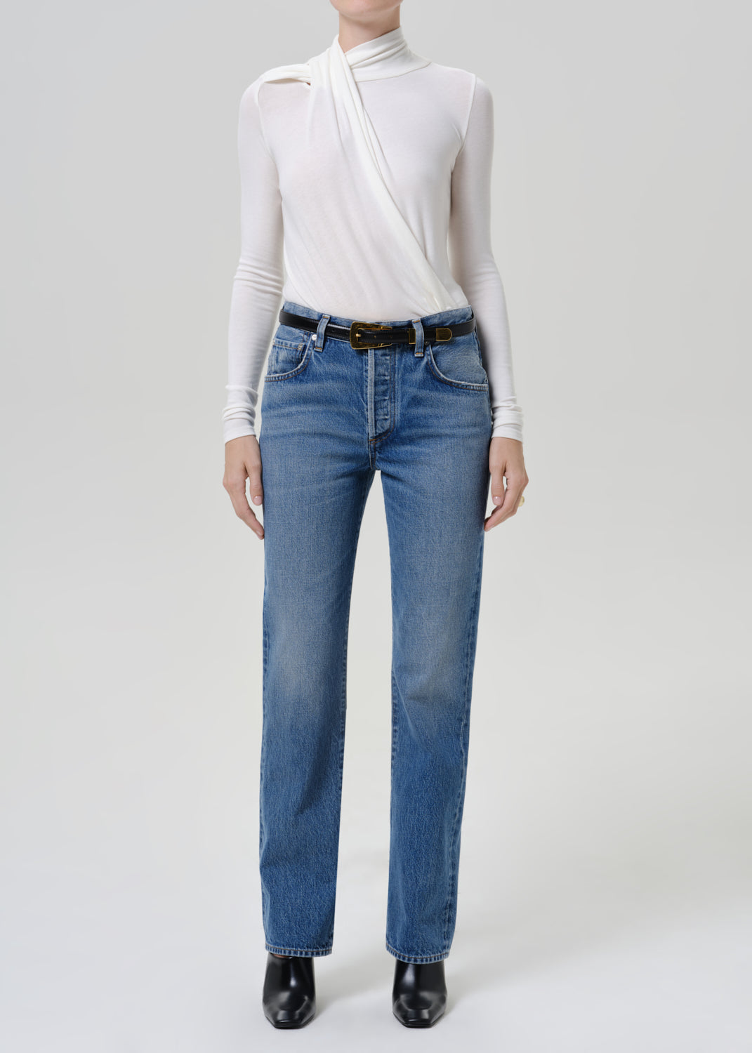 Woman wearing medium blue straight leg jeans with a white long sleeve mock neck top on a grey background.