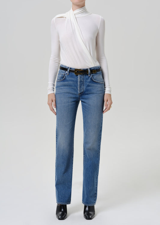Woman wearing medium blue straight leg jeans with a white long sleeve mock neck top on a grey background.