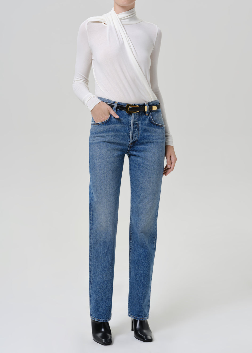 Woman wearing medium blue straight leg jeans with a white long sleeve mock neck top on a grey background.