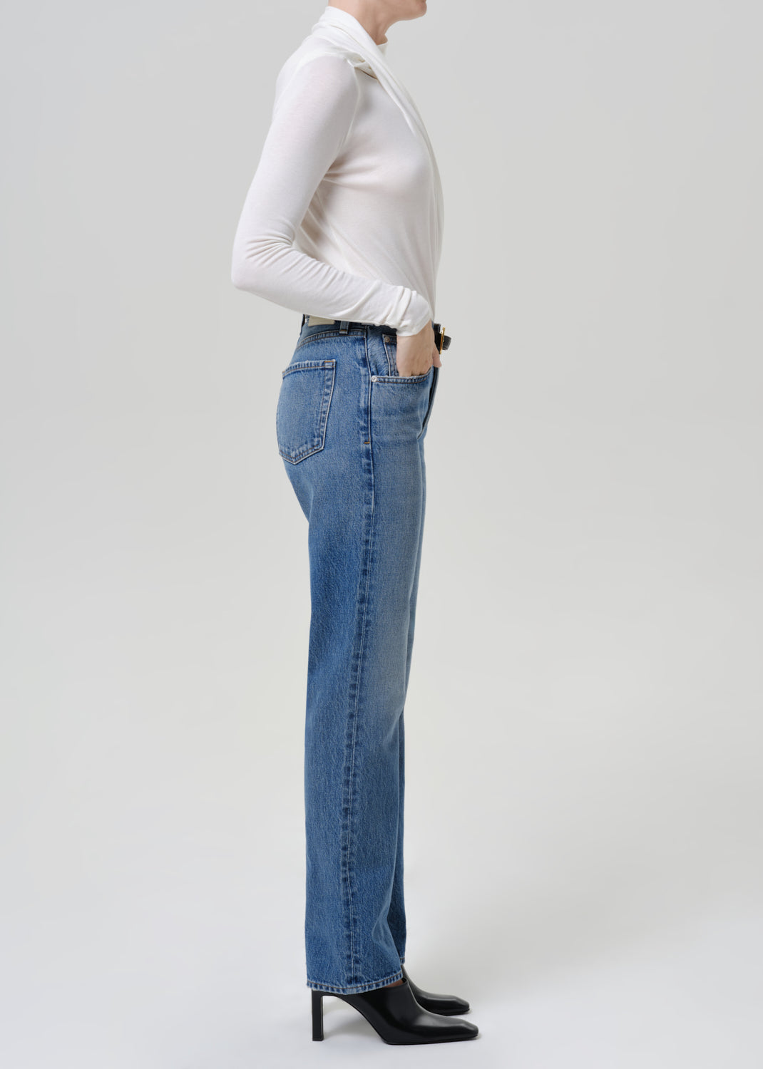 Woman wearing medium blue straight leg jeans with a white long sleeve mock neck top viewed from the side on a grey background.