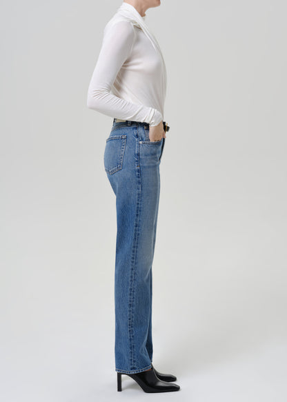 Woman wearing medium blue straight leg jeans with a white long sleeve mock neck top viewed from the side on a grey background.