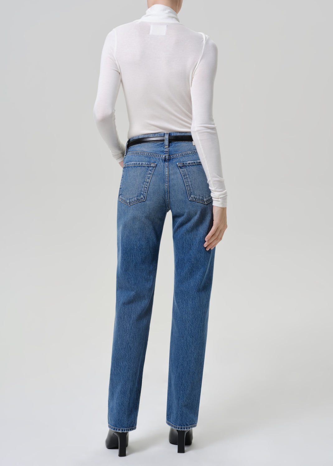 A model shown from the back wearing blue straight-leg jeans, a white long-sleeve turtleneck top, and black heels against a light gray studio background.