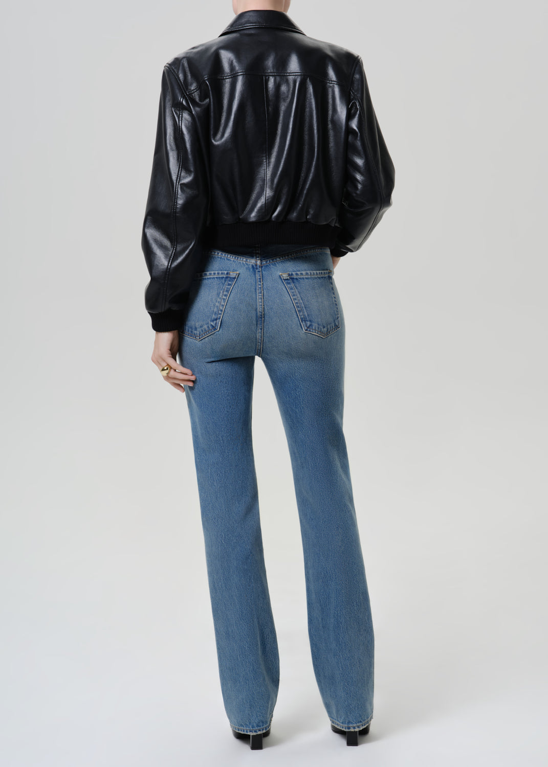 Woman wearing medium blue straight leg jeans with a black leather jacket viewed from the back on a grey background.