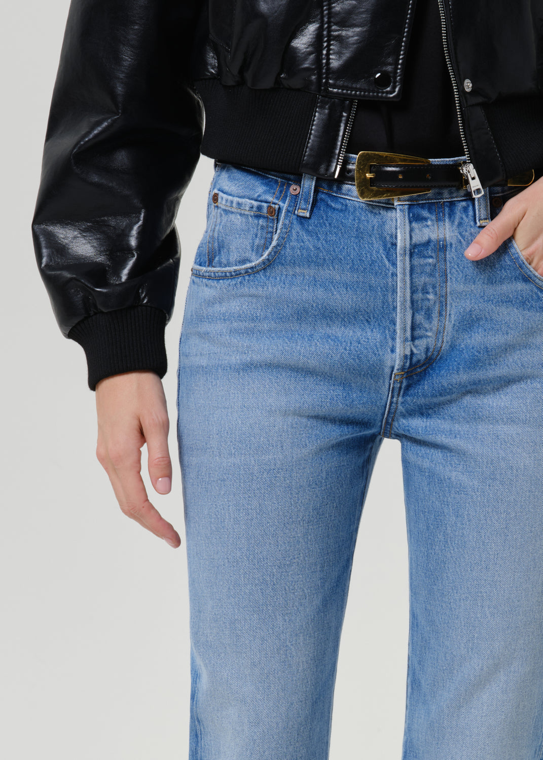 Close-up of a person wearing light blue straight-leg jeans with a black leather jacket and a gold-tone belt, shown against a plain light gray studio background.