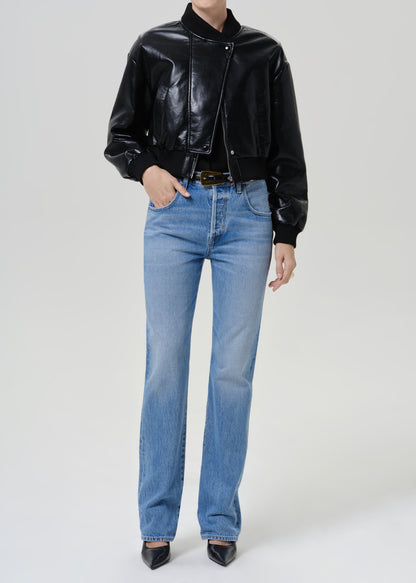 A model wearing a black glossy cropped leather jacket, light wash blue straight-leg jeans, and black pointed-toe heels, standing against a light gray studio background.