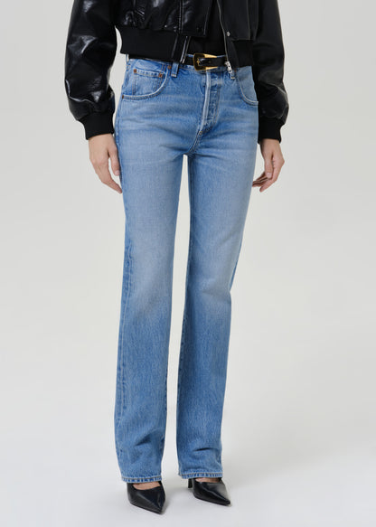 A model wearing a pair of light blue straight-leg denim jeans, styled with a black leather jacket, black belt, and black pointed-toe heels against a light gray studio background.