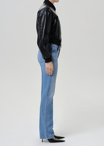 A side-profile studio shot of a model wearing light-wash blue straight-leg jeans, a black leather jacket, and black pointed-toe heels against a light gray background.