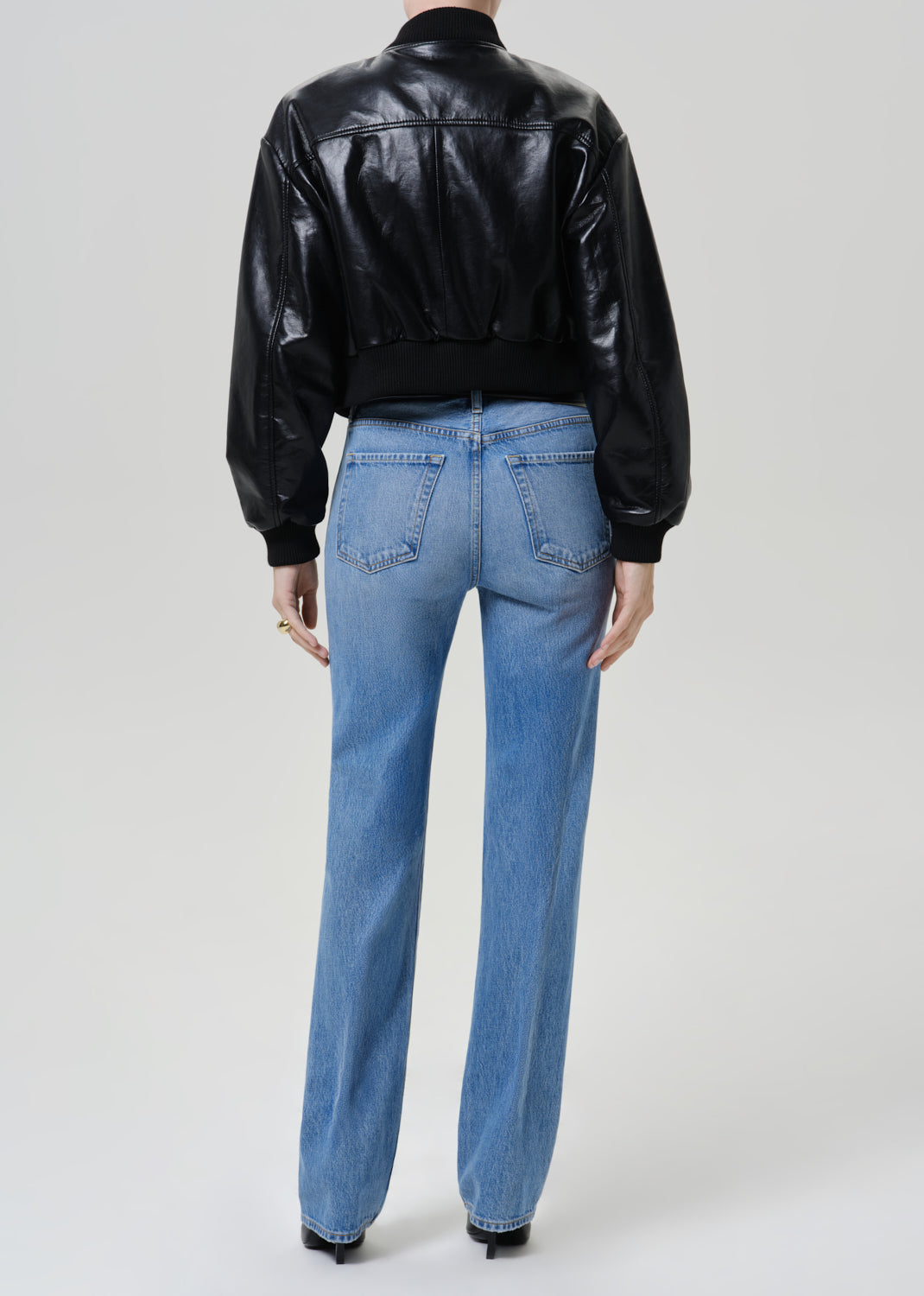 A model shown from the back wearing a black leather cropped bomber jacket and light blue straight-leg jeans, photographed against a neutral light gray studio background.