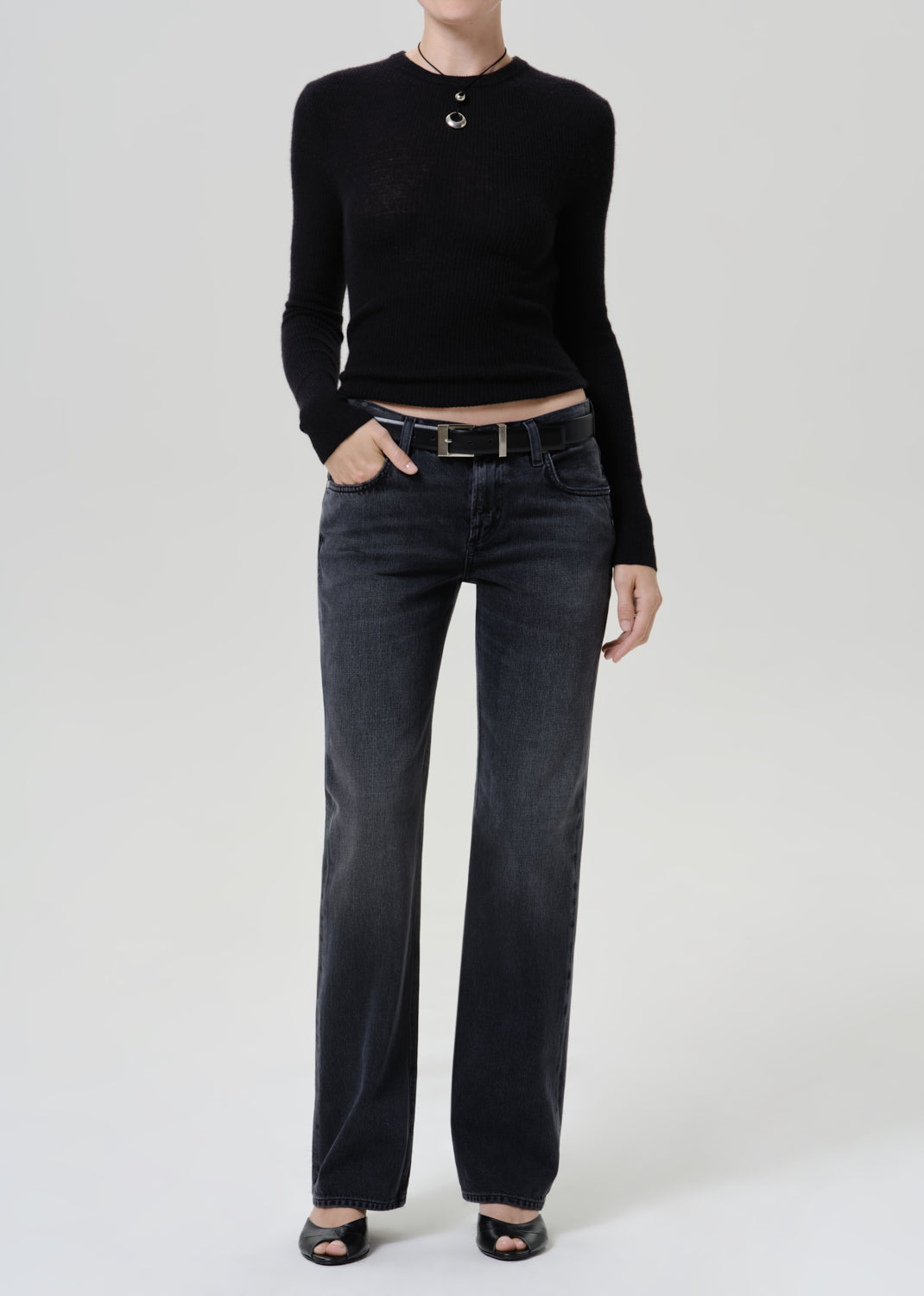 A model wearing a black long-sleeve cropped knit top, dark gray straight-leg jeans with a black belt, and black open-toe heels, standing against a light gray studio background.