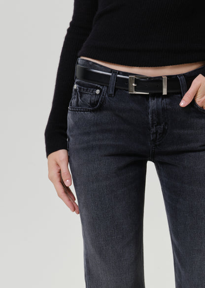 Close-up of a person wearing dark charcoal gray jeans with a black belt featuring a silver rectangular buckle, paired with a black ribbed long-sleeve top against a light gray studio background.
