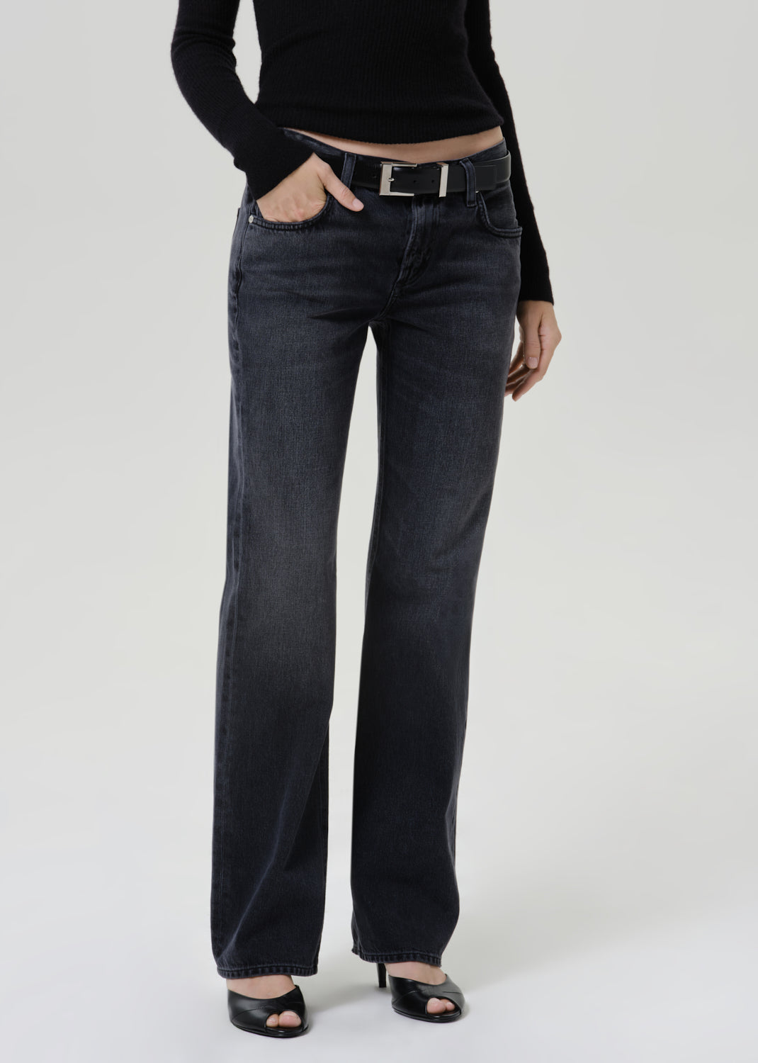 A model wearing a pair of dark charcoal gray wide-leg jeans with a black belt, paired with a black long-sleeve top and black heeled sandals, photographed against a light gray studio background.