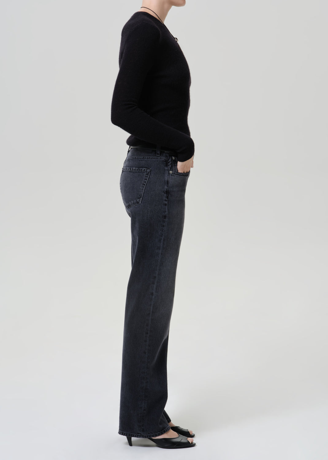 A side-profile studio shot of a model wearing dark charcoal gray wide-leg jeans, a black long-sleeve top, and black pointed-toe heels against a light gray background.
