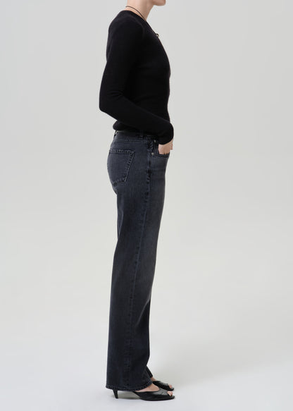 A side-profile studio shot of a model wearing dark charcoal gray wide-leg jeans, a black long-sleeve top, and black pointed-toe heels against a light gray background.