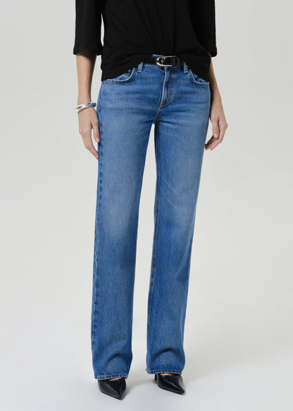 A model wearing a pair of medium-wash blue straight-leg jeans with a black top and black pointed shoes, standing against a light gray studio background.