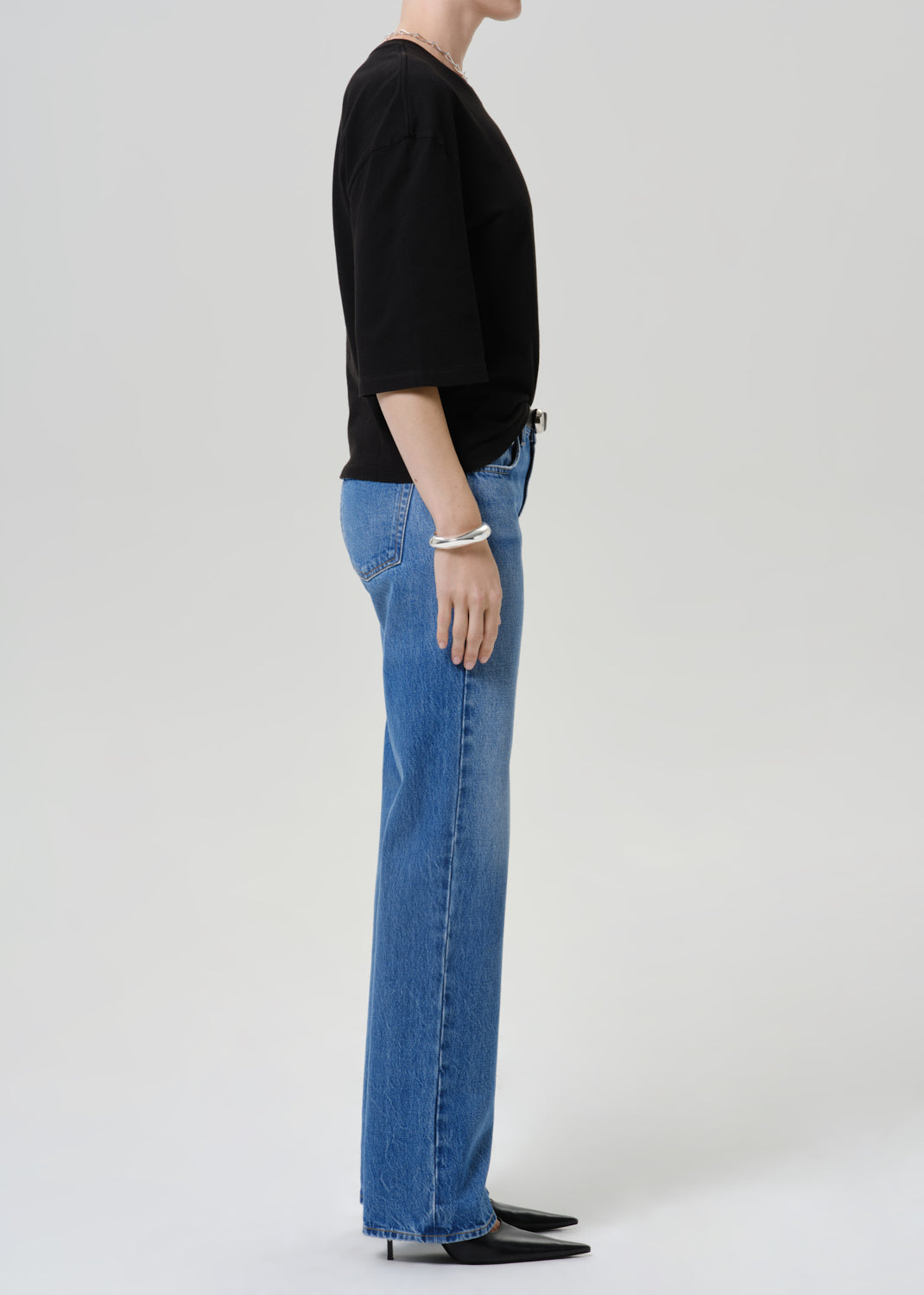 A side-profile view of a model wearing a black short-sleeve top and blue wide-leg jeans against a light gray studio background.