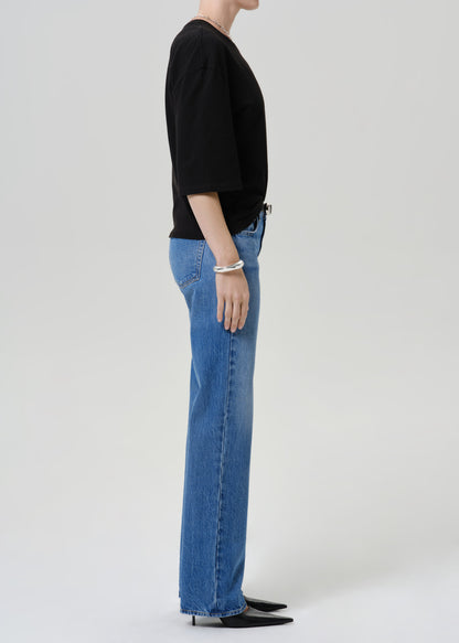 A side-profile view of a model wearing a black short-sleeve top and blue wide-leg jeans against a light gray studio background.