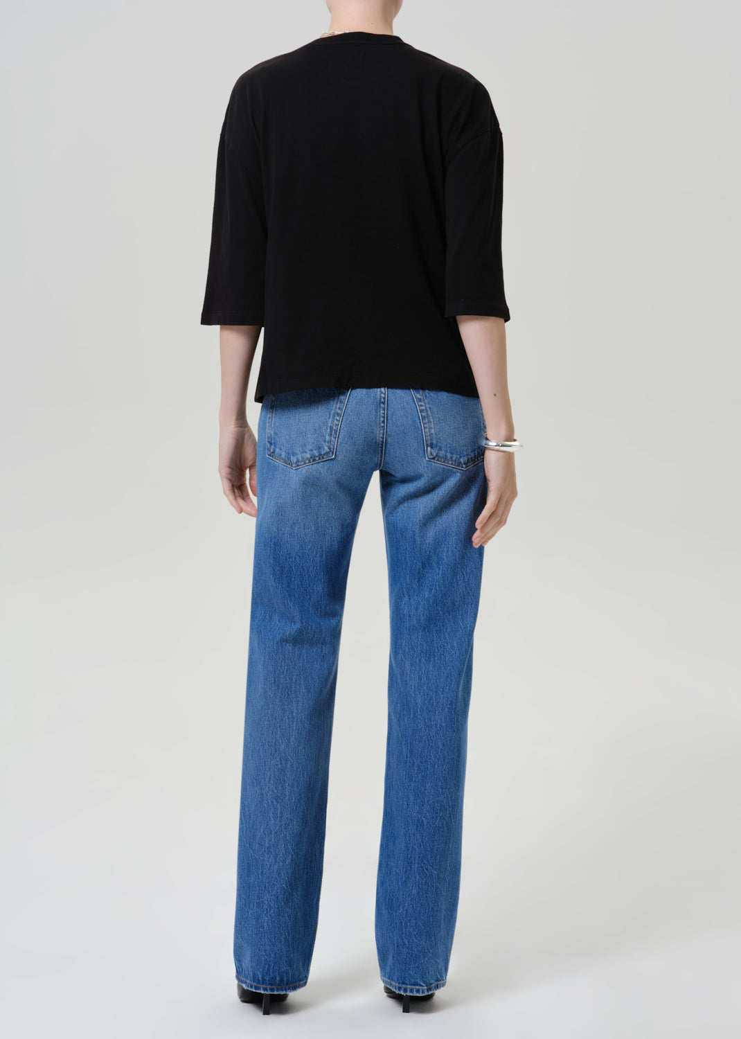A model shown from the back wearing a black three-quarter sleeve top and blue straight-leg jeans against a light gray studio background.